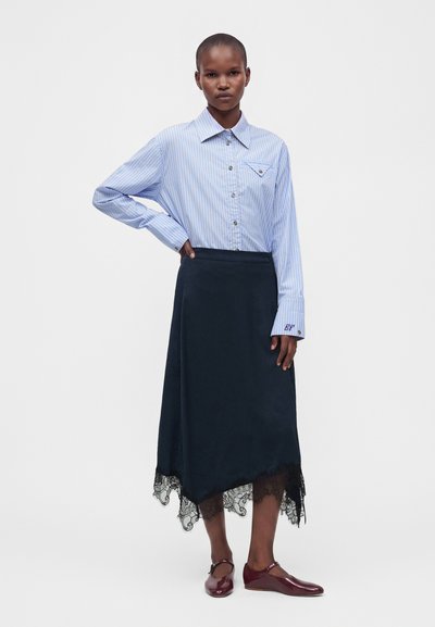 Femme portant une chemise à rayures bleues boutonnée, une jupe midi foncée avec un ourlet en dentelle et des chaussures plates bordeaux, debout, une main sur la hanche.