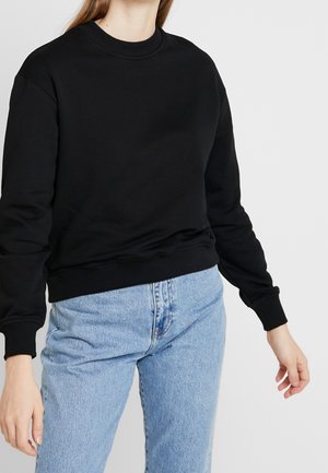 Kobieta w czarnej bluzie z długim rękawem i okrągłym dekoltem oraz jasnoniebieskich, wysokich spodniach dżinsowych na jednolitym tle.