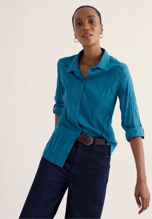 Femme portant une chemise turquoise à boutons avec manches retroussées, un jean foncé et une ceinture marron, debout devant un fond clair.