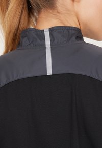 Svart sportjacka med en grå, reflexiv yoke-detalj och ståkrage, tillverkad av lätt, strukturerad tyg.