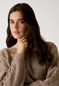 Brązowy sweter dzianinowy z prążkowanymi mankietami i teksturowanymi akcentami. Modelka pozuje z ręką przy szyi, prezentując miękki, przytulny materiał.