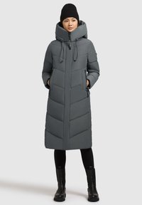 Grå, knælang puffjakke med høj krave, lynlåslukning, sidelommer og chevron-stikninger. Bæret over sorte leggings og støvler.