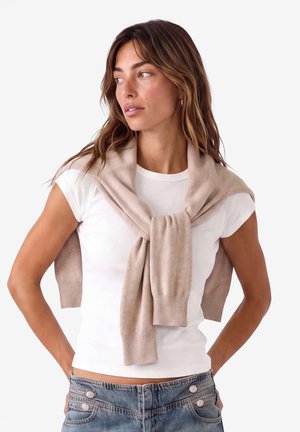 Kvinde med langt brunt hår iført en hvid t-shirt og en beige sweater draperet over skuldrene, kigger til siden med hænderne på hoften.