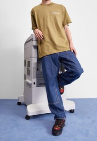 Camiseta de algodón marrón con texto blanco, combinada con pantalones de trabajo de mezclilla azules y zapatos negros con detalles en cuadros rojos, apoyado contra una impresora.