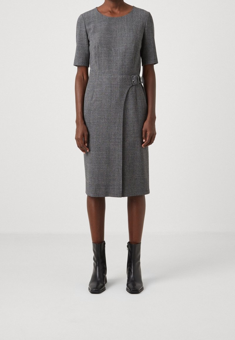 Femme portant une robe à carreaux grise longueur genou, manches courtes, avec un détail de boucle sur le côté et des bottines noires.
