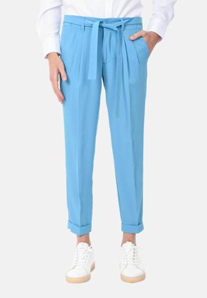 Pantaloni su misura blu chiaro con vita legata, pieghe, tasche laterali e orli arrotolati. Abbinati a scarpe da ginnastica bianche su uno sfondo neutro.