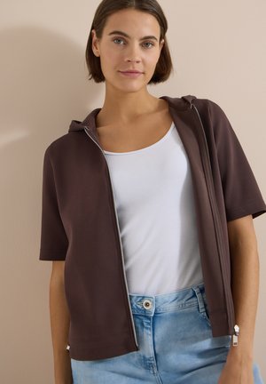 Jeune femme aux cheveux courts et bruns portant un débardeur blanc, un sweat à capuche marron à manches courtes avec fermeture éclair et un jean bleu clair devant un mur beige.