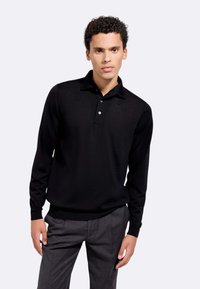 Polo a maniche lunghe nera realizzata in tessuto a maglia, con colletto e chiusura a tre bottoni. Indossata con pantaloni grigi su misura.