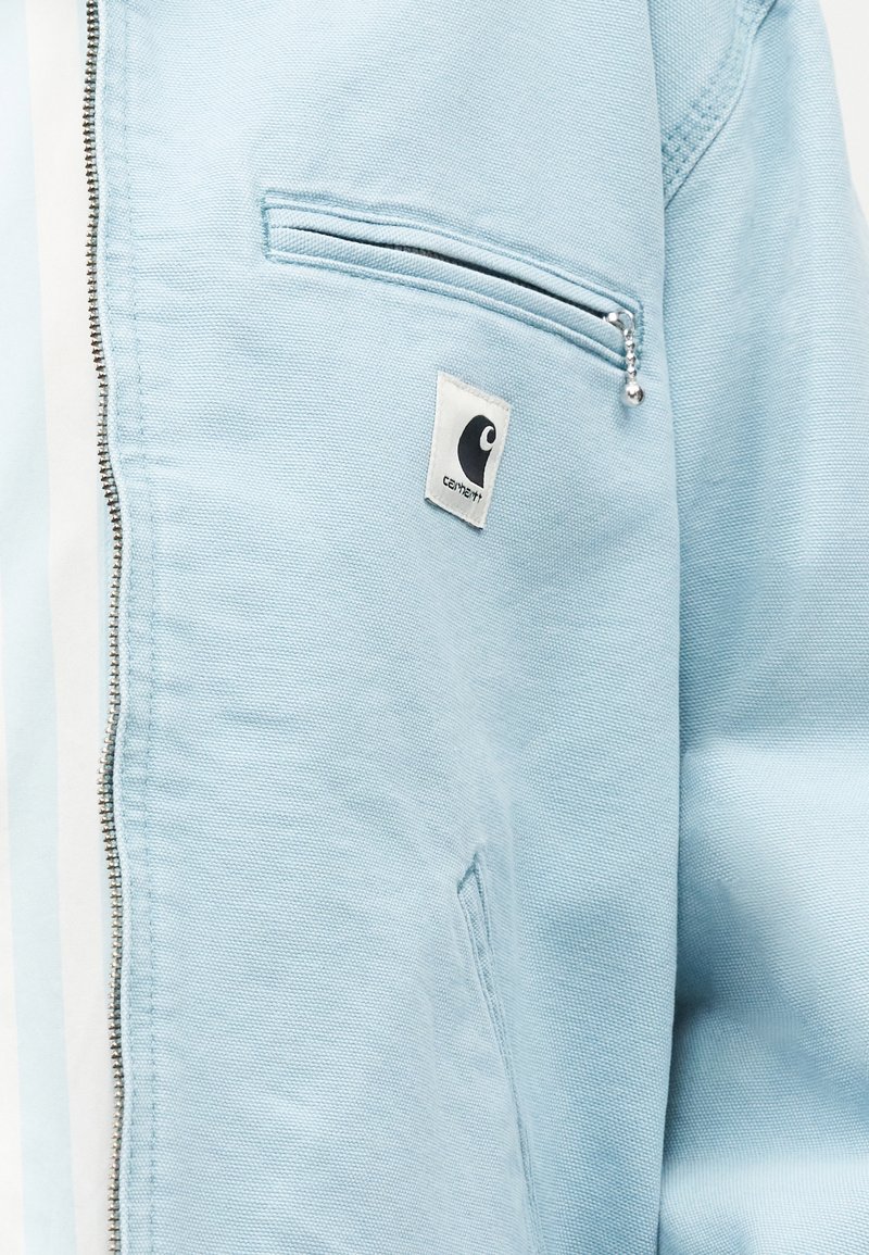 Veste bleu clair avec une fermeture éclair métallique et une poche poitrine zippée ornée d'un petit patch logo de marque rectangulaire.