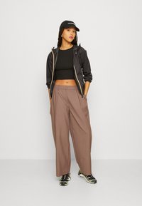 Zwarte hoodie met gouden rits, zwarte cropped top, bruine wijde broek en zwarte sneakers met groene accenten. Een zwarte pet maakt de outfit af.