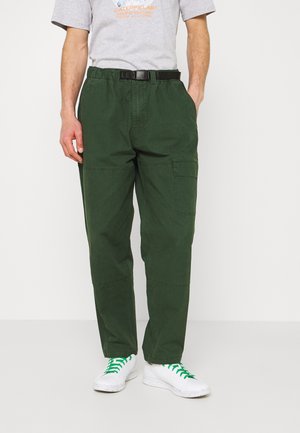 Man draagt donker groene cargobroek met een zwarte riem, een grijs Caterpillar T-shirt en witte sneakers met groene veters, staand met handen in de zakken.