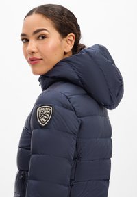 Frau mit zurückgebundenen Haaren, die eine marineblaue Steppjacke mit Kapuze und einem Schild-Ärmelabzeichen trägt und über ihre Schulter blickt.