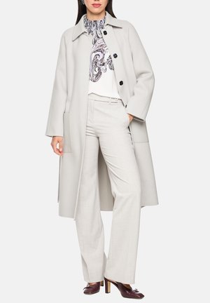 Woman wearing a light gray long coat over patterned blouse, matching trousers, and dark brown high-heeled shoes, standing with one hand in pocket.