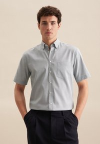Camicia a maniche corte con bottoni in tessuto grigio chiaro, dotata di taschino sul petto, colletto classico e una fine trama. Indossata con pantaloni a pieghe scuri.