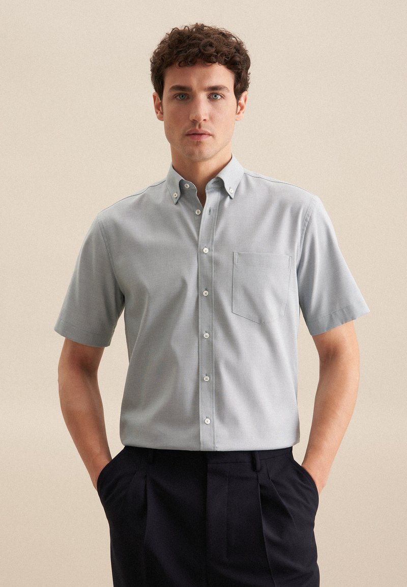 Camicia a maniche corte con bottoni in tessuto grigio chiaro, dotata di taschino sul petto, colletto classico e una fine trama. Indossata con pantaloni a pieghe scuri.