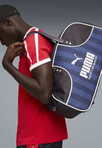 Person iført en rød- og hvidstribet skjorte med en sort kasket, bærer en stor blå og sort Puma rygsæk over den ene skulder.