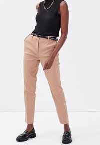Pantalons en toile beige ajustés avec des plis avant, associés à un haut sans manches noir en côtes et des mocassins noirs avec des ornements en clous.