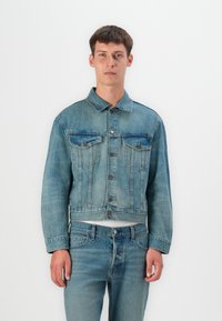 Denim jacket in light blue with a classic cut, two chest pockets, metal buttons, and subtle fading. Worn with matching jeans.