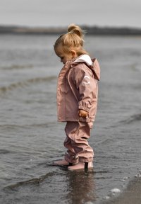 Roza vodoodporna jakna in hlače z elastičnimi manšetami in kapuco. Stoje v plitvi vodi, obut v ujemajoče se roza škornje s črnimi poudarki.