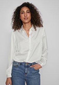 Camicia bianca con bottoni in tessuto liscio, colletto, maniche lunghe e una vestibilità rilassata, abbinata a jeans blu a vita alta.