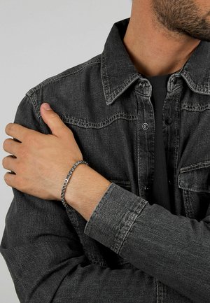 Bracciale in argento con maglie piatte indossato su una mano, in contrasto con una giacca di jeans grigio scuro con bottoni e tessuto testurizzato.