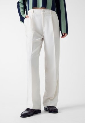 Persona che indossa pantaloni bianchi su misura a vita alta con pieghe, abbinati a mocassini neri e a un maglione corto a righe blu navy e verde.