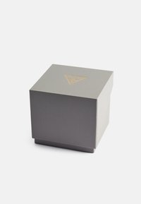 Caja gris con una superficie texturizada, que presenta un logo triangulo dorado en la parte superior. Forma cuadrada con una ligera indentación en un lado.
