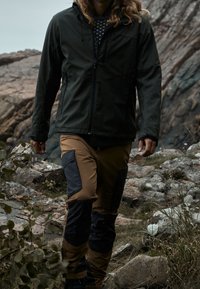 Osoba ubrana w czarną kurtkę i brązowe spodnie z czarnymi łatami, id�ąca po skalistym terenie z roślinnością.