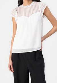 Žena v bílém bezramínkovém topu s krajkovým lemem a volánkovými rukávy, doplněná černými kalhotami s vysokým pasem.