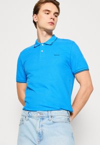 Cămașă polo din bumbac albastru cu un design clasic, având guler cu nasturi și accente negre pe mâneci. Logo pe piept.