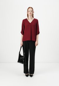 Burgunderfarbige V-Ausschnitt-Bluse mit 3/4 gerollten Ärmeln, kombiniert mit schwarzen, taillierten Hosen und spitzen schwarzen Schuhen, dabei eine schwarze Lederhandtasche haltend.