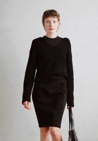 Pull en tricot noir avec un col en V et une texture côtelée, superposé à une jupe crayon noire. Le modèle tient un sac fourre-tout en cuir noir.