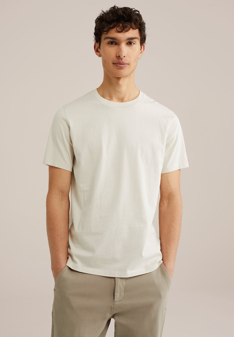 Tricou scurt cu mânecă scurtă, de culoare bej deschis, din bumbac, cu decolteu rotund și textură netedă, purtat cu pantaloni de culori deschise.