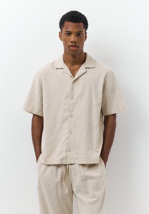 Nuori mies, jolla on päällään löysä beige lyhythihainen nappipaita ja siihen sopivat narulla kiristetyt housut, seisomassa käsien ollessa taskuissa valkoista taustaa vasten.