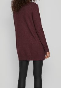 Burgundy knitted cardigan featuring a long, relaxed fit, ribbed cuffs, and a round hem, paired with black fitted leggings.