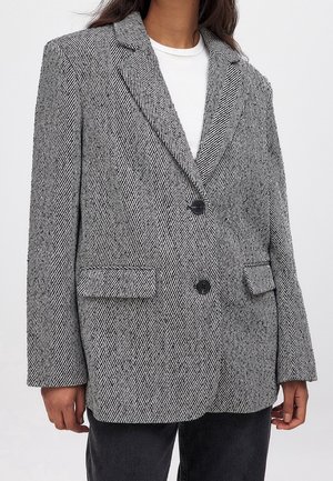 Vrouw draagt een zwart-witte visgraatblazer met twee knopen en klepzakken over een wit topje en zwarte broek.