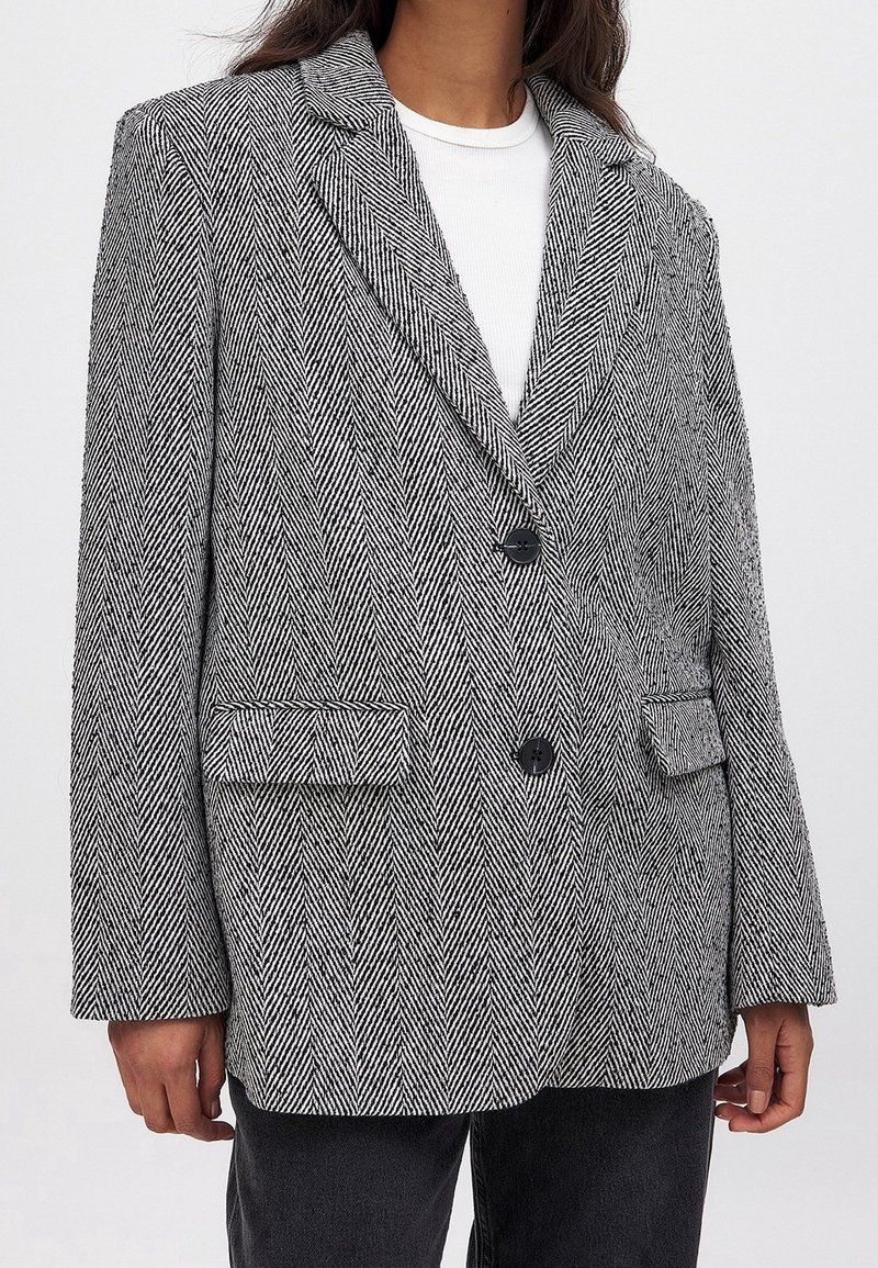 Femme portant un blazer à chevrons noir et blanc avec deux boutons et des poches à rabat, sur un haut blanc et un pantalon noir.