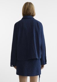 Giacca navy, testurizzata, con una vestibilità comoda e maniche lunghe. Presenta un colletto e dettagli a motivi sottili. Abbinata a una gonna coordinata.