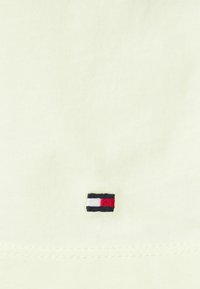 Tissu en coton jaune clair avec un petit logo brodé comportant des rayures horizontales rouges, blanches et noires dans le coin inférieur.