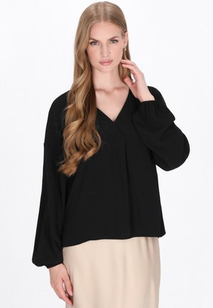 Blusa nera ampia con scollatura a V, maniche lunghe a palloncino, tessuto morbido e dettagli di pieghe sottili sulla parte anteriore, indossata su un indumento beige.