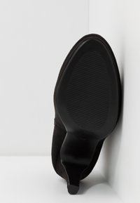 Zapato de tacón alto de ante negro con punta afilada y suela de goma texturizada, exhibido sobre un fondo blanco, destacando su forma curvada.