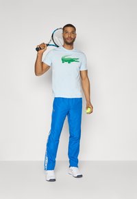 Lichtblauw T-shirt met groen krokodillenlogo, gecombineerd met felblauwe trainingsbroek met witte zijstrepen. Houdt een tennisracket en bal vast.