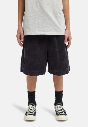 Persona con pantaloncini neri di velluto a coste fino al ginocchio, calze nere, scarpe da ginnastica nere e bianche e una maglietta grigio chiaro, in piedi contro uno sfondo bianco.