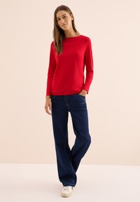 Röd långärmad t-shirt i mjukt tyg, kombinerad med mörkblå vida jeans. Skorna är vita med en minimalistisk design. Ljus bakgrund.