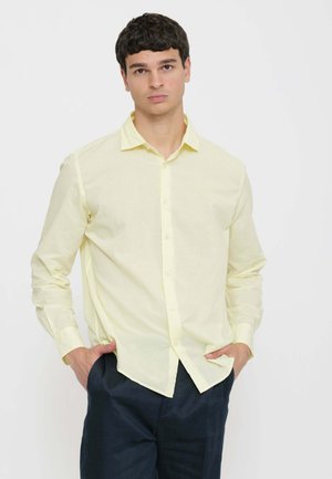 Giovane uomo con capelli corti e scuri che indossa una camicia giallo pallido con bottoni e pantaloni scuri, posando con le mani nelle tasche davanti a uno sfondo semplice.