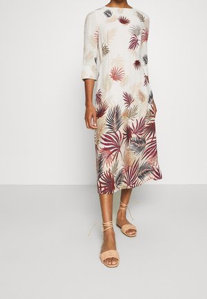 Kvinde iført en hvid midi-kjole med beige, burgundy og sort tropisk bladprint, 3/4-ærmer og beige snøresandaler.