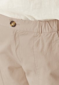 Beige trousers featuring an elastic waistband and a single decorative button. Fabric has a smooth texture with subtle stitching details.