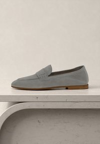 Mocassin en daim gris clair avec une semelle en bois et un petit détail de bride embossé du nom de la marque, posé sur une surface en pierre claire.