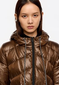 Brązowa, pikowana kurtka puchowa z kapturem, z czarnym zamkiem błyskawicznym i troczkami. Gładka tekstura z połyskującym wykończeniem.