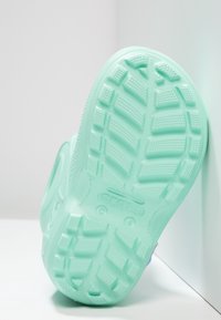 Semelle de chaussure en caoutchouc vert menthe clair avec un motif de semelle texturé, présentant des bosses et des rainures surélevées pour une meilleure traction. Logo de la marque visible.
